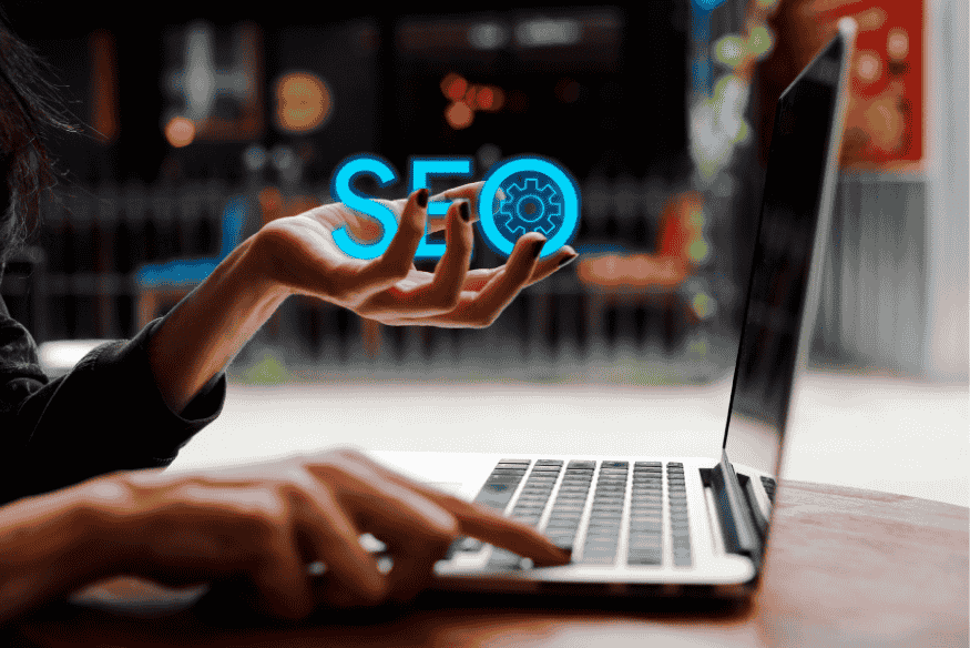 Mão segurando ícone digital de SEO em frente a um notebook, representando controle estratégico de otimização.