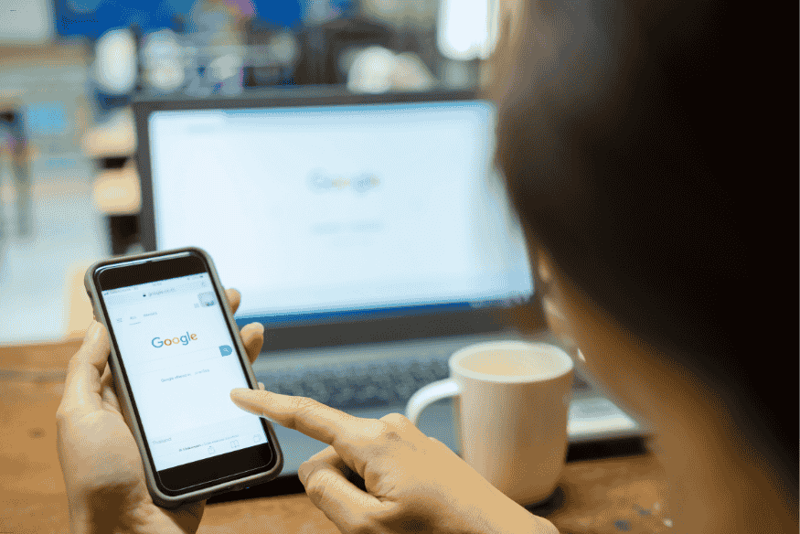 Pessoa usando celular com Google aberto, com notebook ao fundo mostrando a mesma busca.